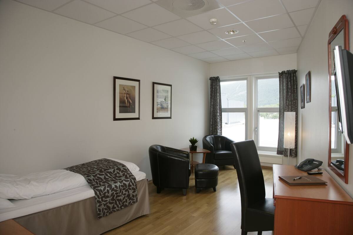 Ein Hotelzimmer mit einem Bett, einem Schreibtisch mit Telefon und Notizbuch, einem Stuhl und zwei gerahmten Bildern an der Wand.
