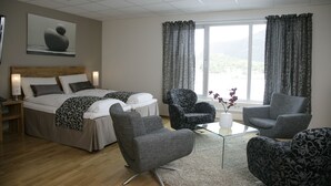 A modern hotel room with a bed, two armchairs, a glass coffee table, and a view of the outdoors.