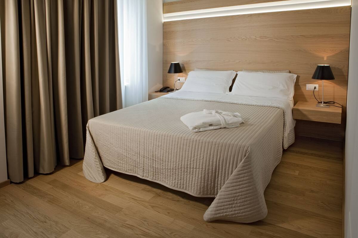 Une chambre d’hôtel avec un grand lit, deux tables de chevet avec des lampes, une tête de lit en bois et une vue sur l’extérieur à travers des rideaux.