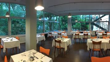Un restaurant avec de nombreuses tables dressées pour le service, de grandes fenêtres donnant sur la verdure et des luminaires suspendus.