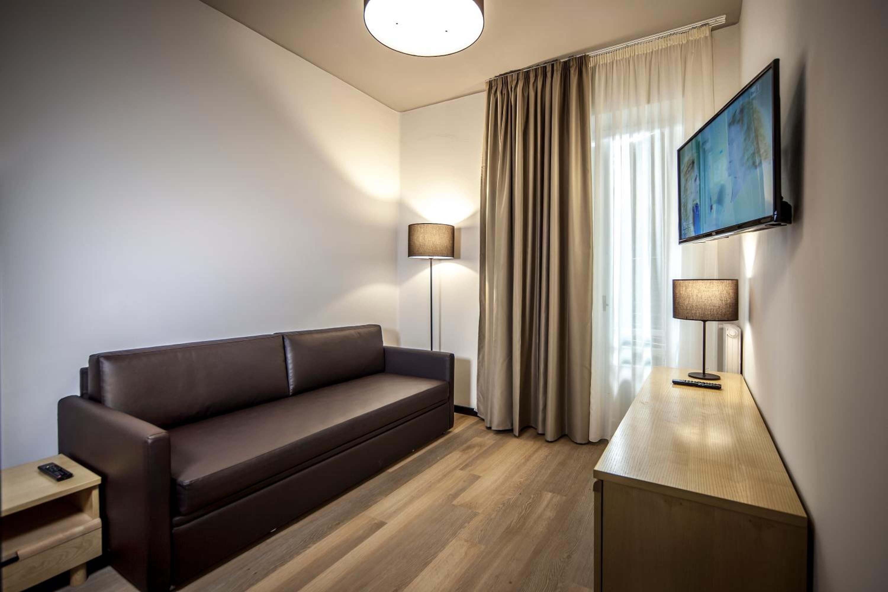 A modern hotel room with a leather sofa, two bedside lamps, a flat-screen TV mounted on the wall, and a large window with curtains.