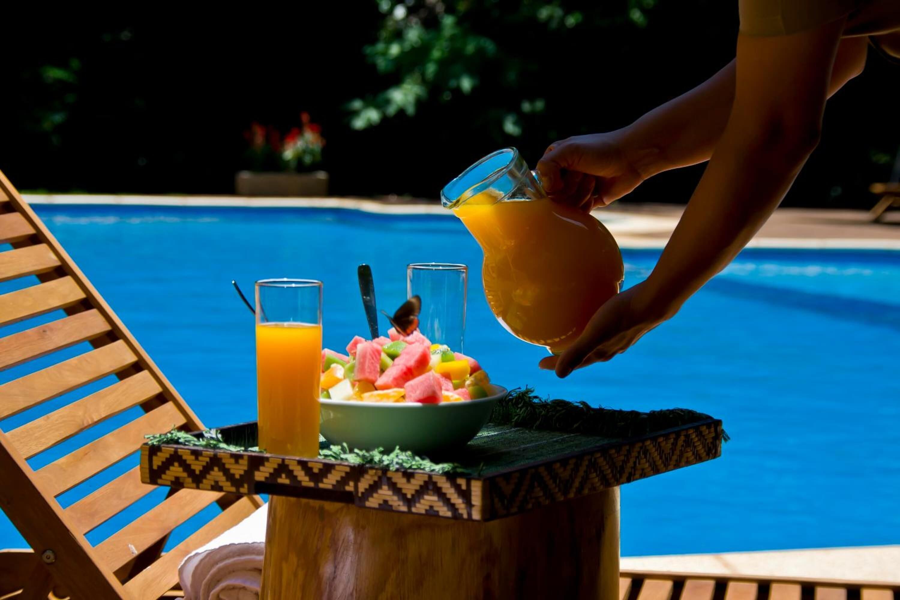 Un décor en bord de piscine avec une chaise en bois, une table sur laquelle se trouve un saladier rempli de fruits et un verre de jus d’orange.
