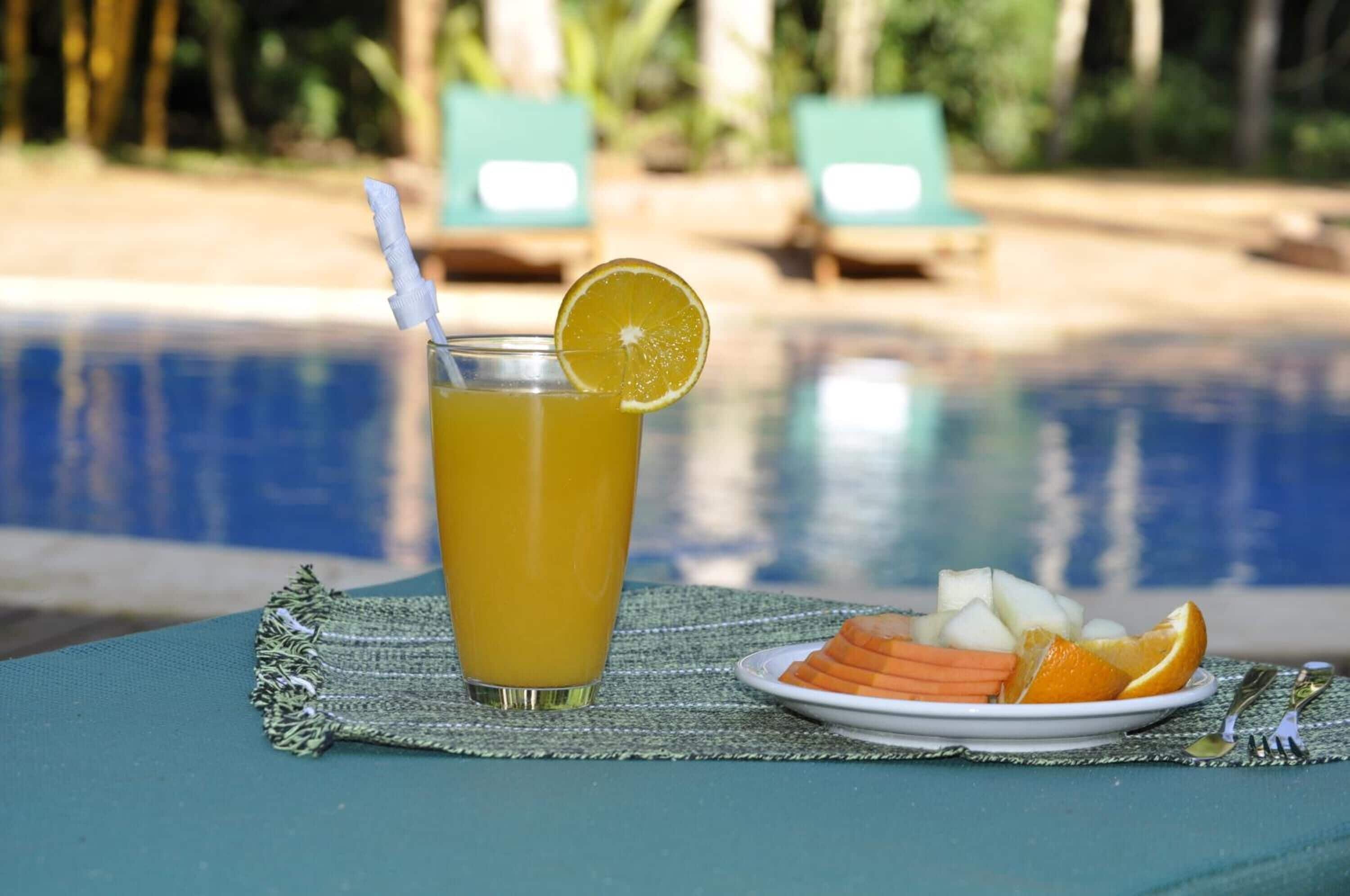 Un vaso de jugo de naranja con una rodaja de limón sobre una mesa, cerca de una piscina con sillones de descanso al fondo.