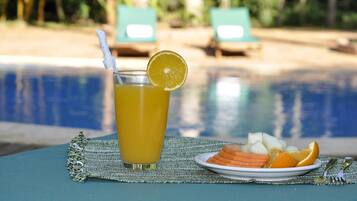 Un vaso de jugo de naranja con una rodaja de limón sobre una mesa, cerca de una piscina con sillones de descanso al fondo.