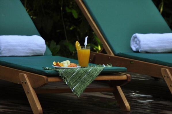 Una silla de lounge de madera con un cojín verde, una toalla blanca doblada, un vaso de jugo de naranja y un plato con fruta y una servilleta.