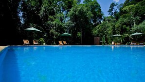 A clear blue pool surrounded by lounge chairs and umbrellas, with dense trees in the background.