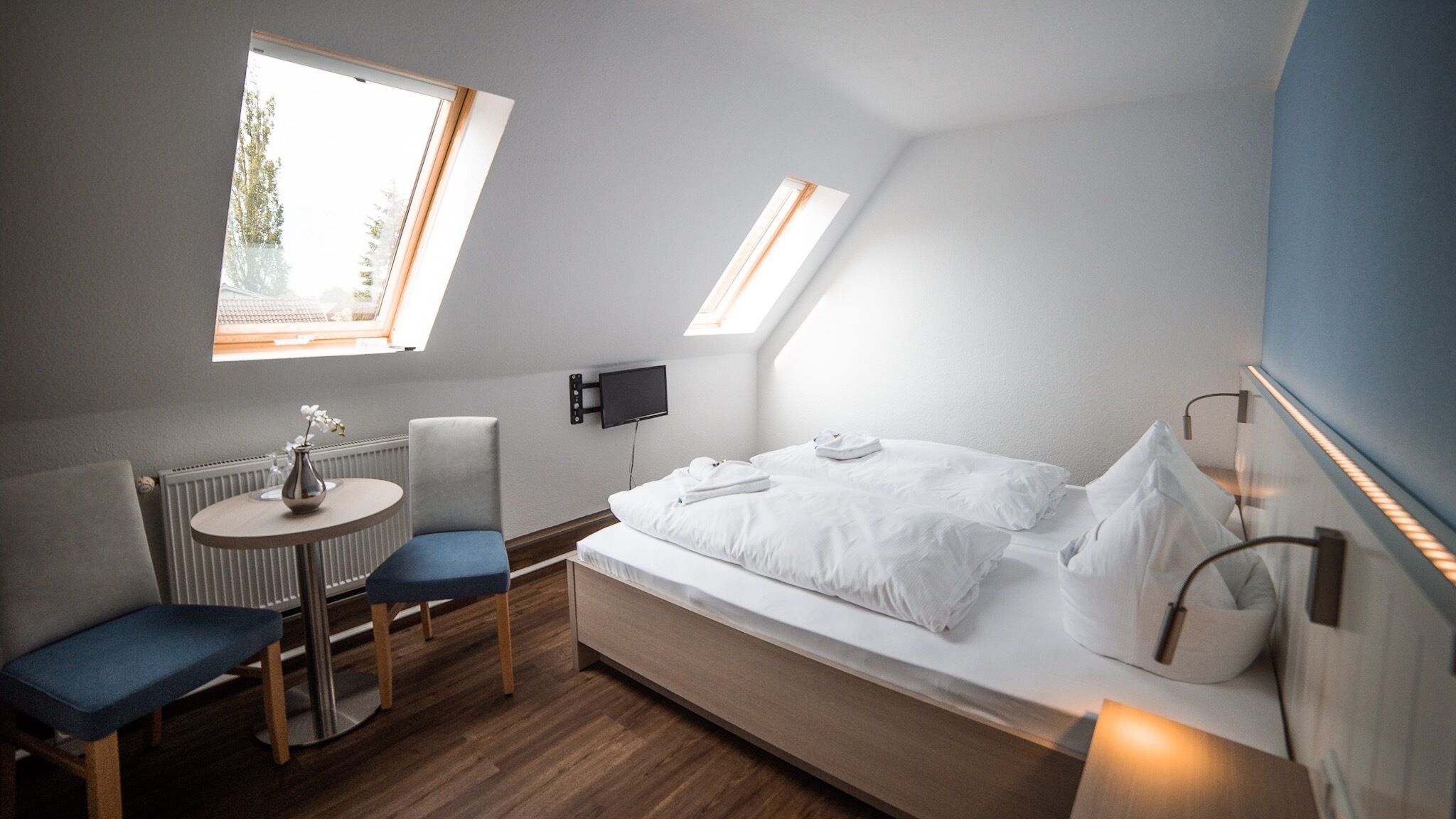 Une chambre compacte et moderne, équipée d’un lit, d’une petite table avec une chaise, d’un radiateur et d’un téléviseur fixé au mur.