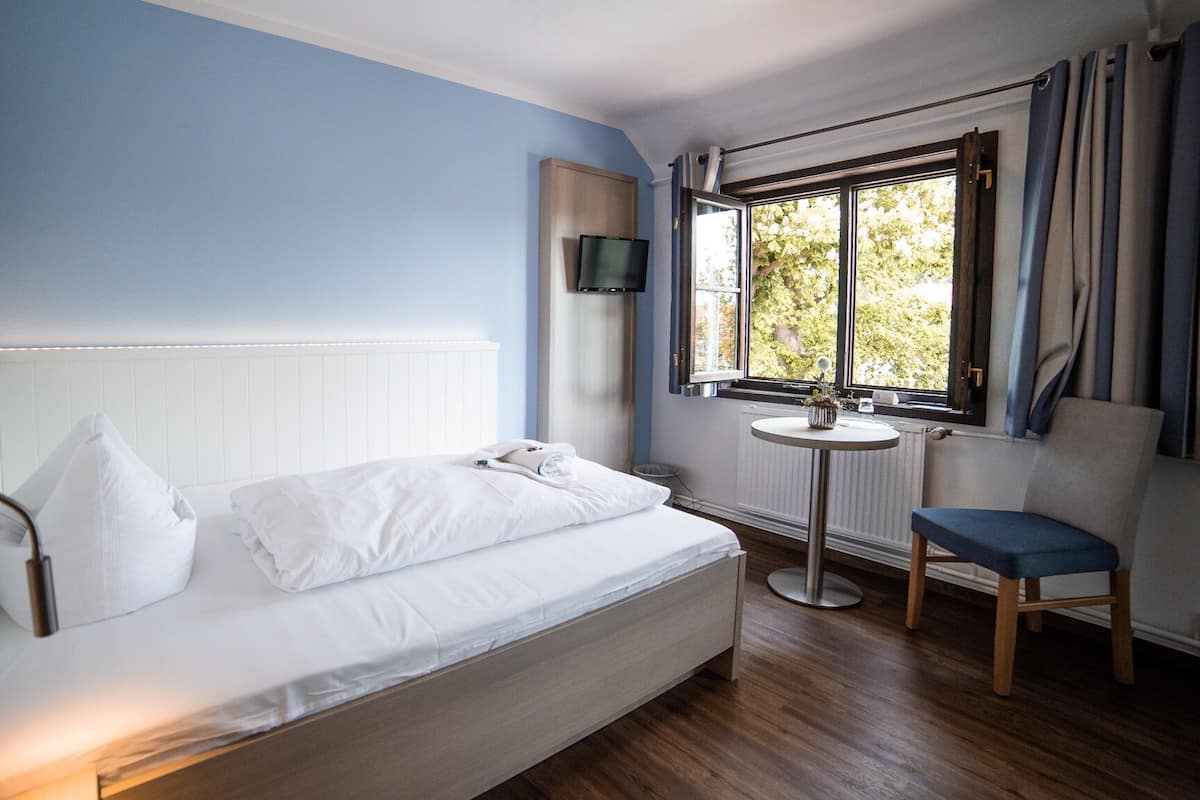 A single bed with white bedding, a bedside table with a plant, a chair, and a TV mounted on the wall.