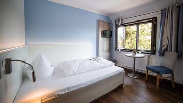 Una cama individual con ropa de cama blanca, una mesita de noche con una planta, una silla y un televisor montado en la pared.