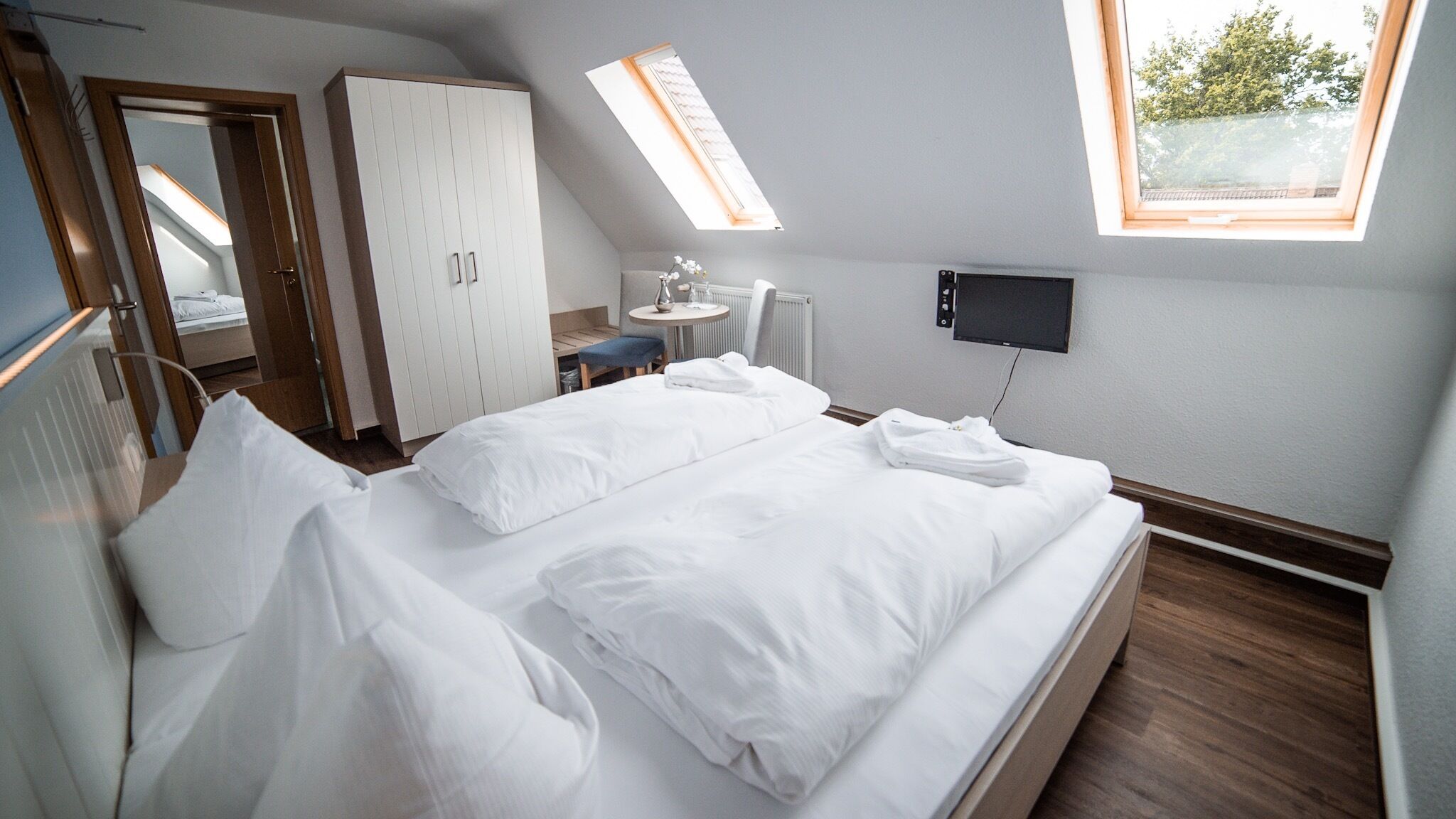 A hotel room with a large bed, a wardrobe, a desk with a chair, a small table, a TV, and a window with a view of trees.