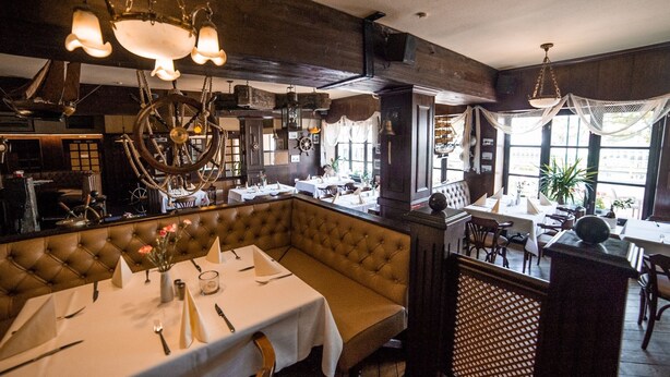 A restaurant with a high ceiling, exposed beams, and a central chandelier. Tables are set with white tablecloths and silverware. - Hotel zur Fähre (Greifswald)