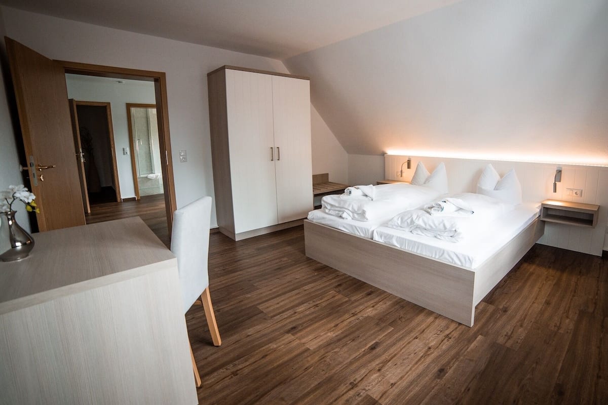 A modern hotel room with a large bed, wooden flooring, a wardrobe, and a desk with a vase of flowers.