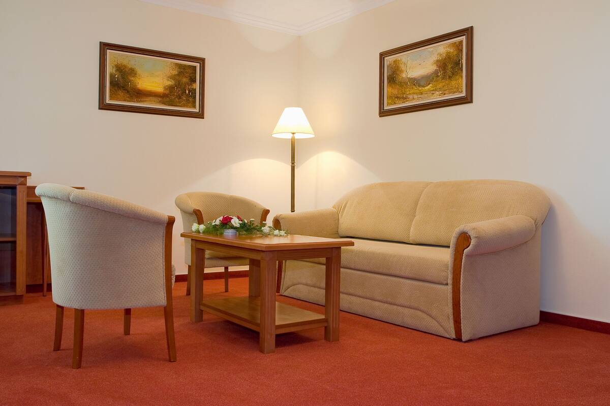 Une pièce chaleureuse avec un canapé beige, un fauteuil blanc et une table basse en bois agrémentée d’un arrangement floral. Deux tableaux encadrés ornent les murs.