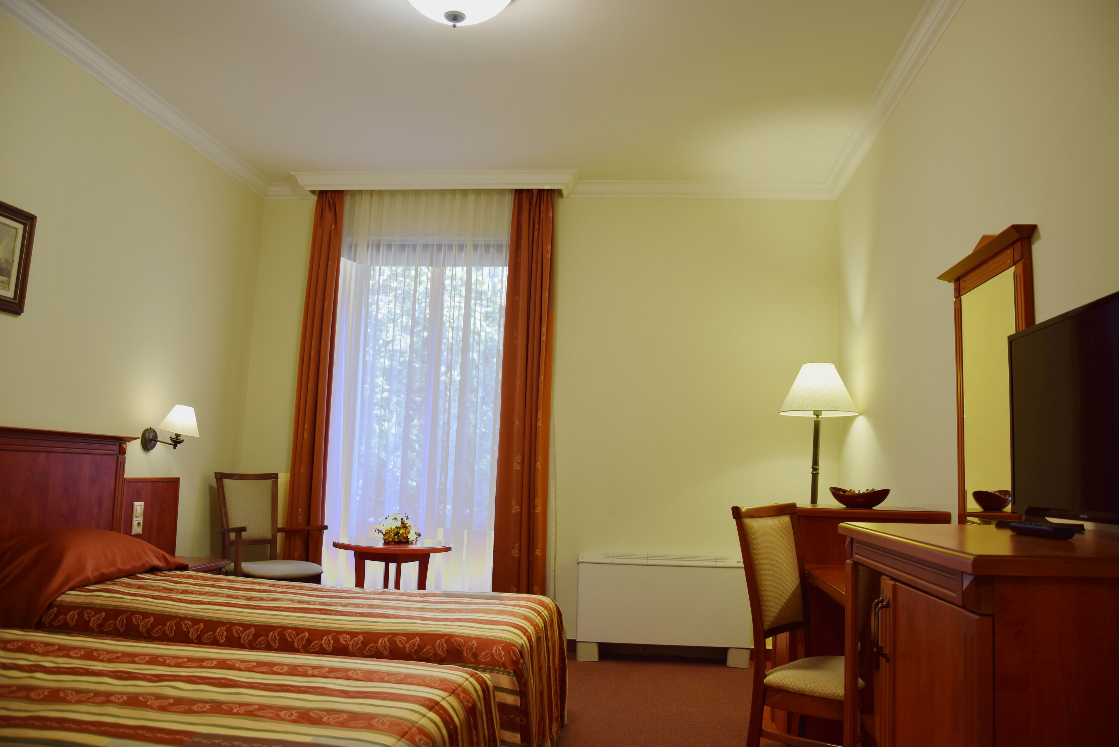 Hotelkamer met twee bedden, een bureau, een stoel, een lamp, een televisie en een raam met gordijnen.