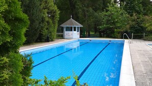 A rectangular swimming pool with clear blue water, surrounded by lush greenery and a gazebo-like structure in the background.