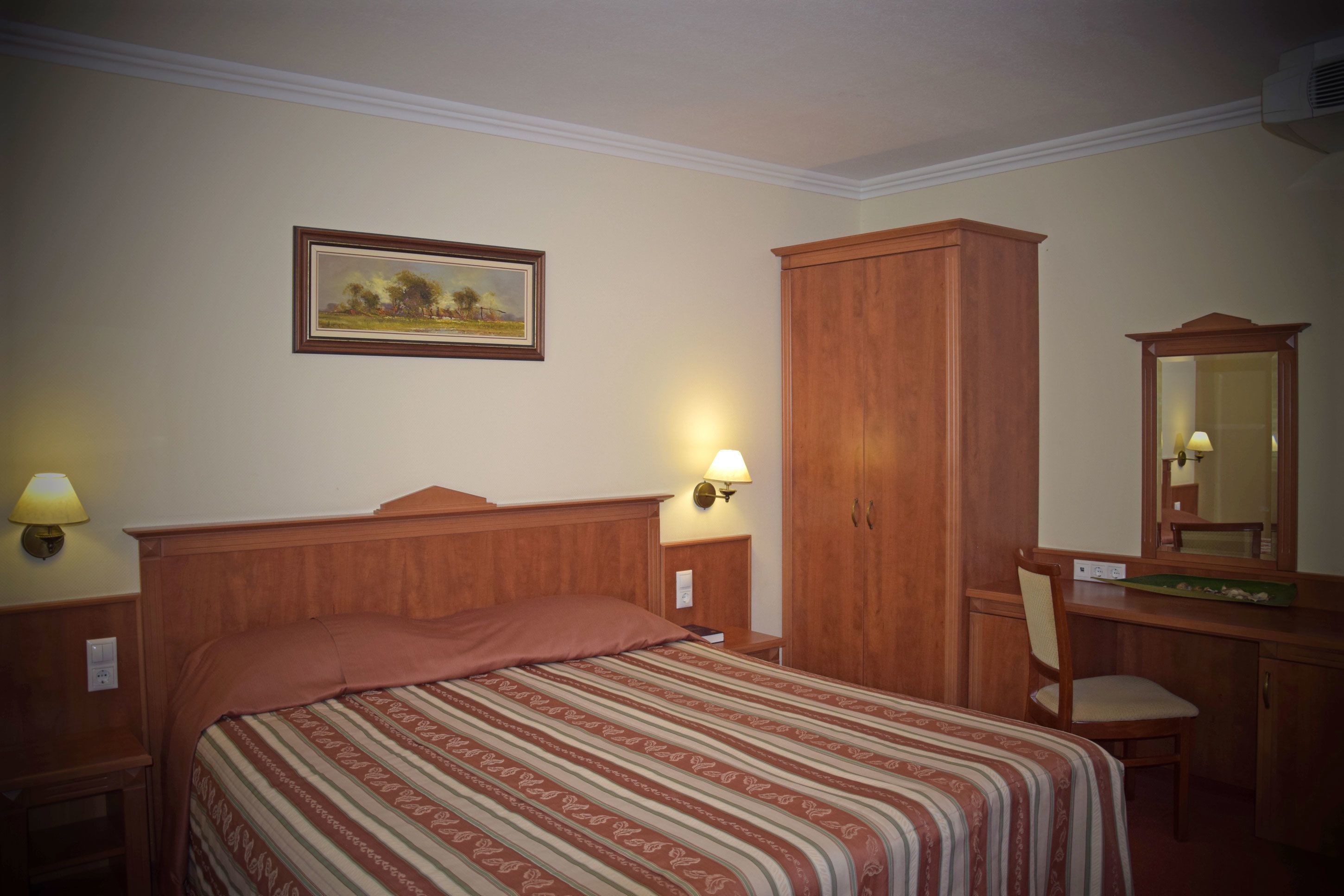 Una habitación de hotel con cama, escritorio, silla, armario, lámpara y un cuadro en la pared.