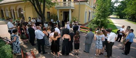A group of people gathered outside a yellow building with a red roof and a balcony.