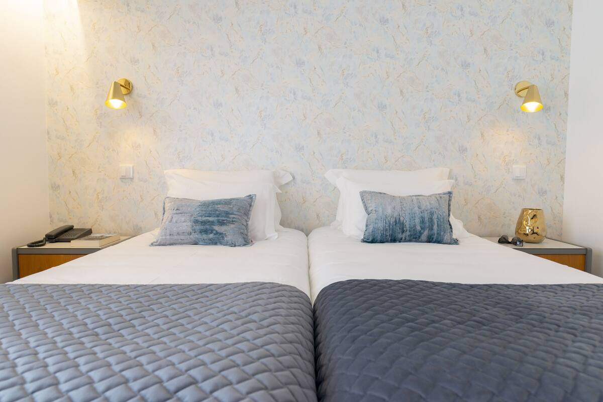 A double bed with a quilted grey bedspread, two pillows, a bedside table with a lamp, and a wall-mounted light fixture.
