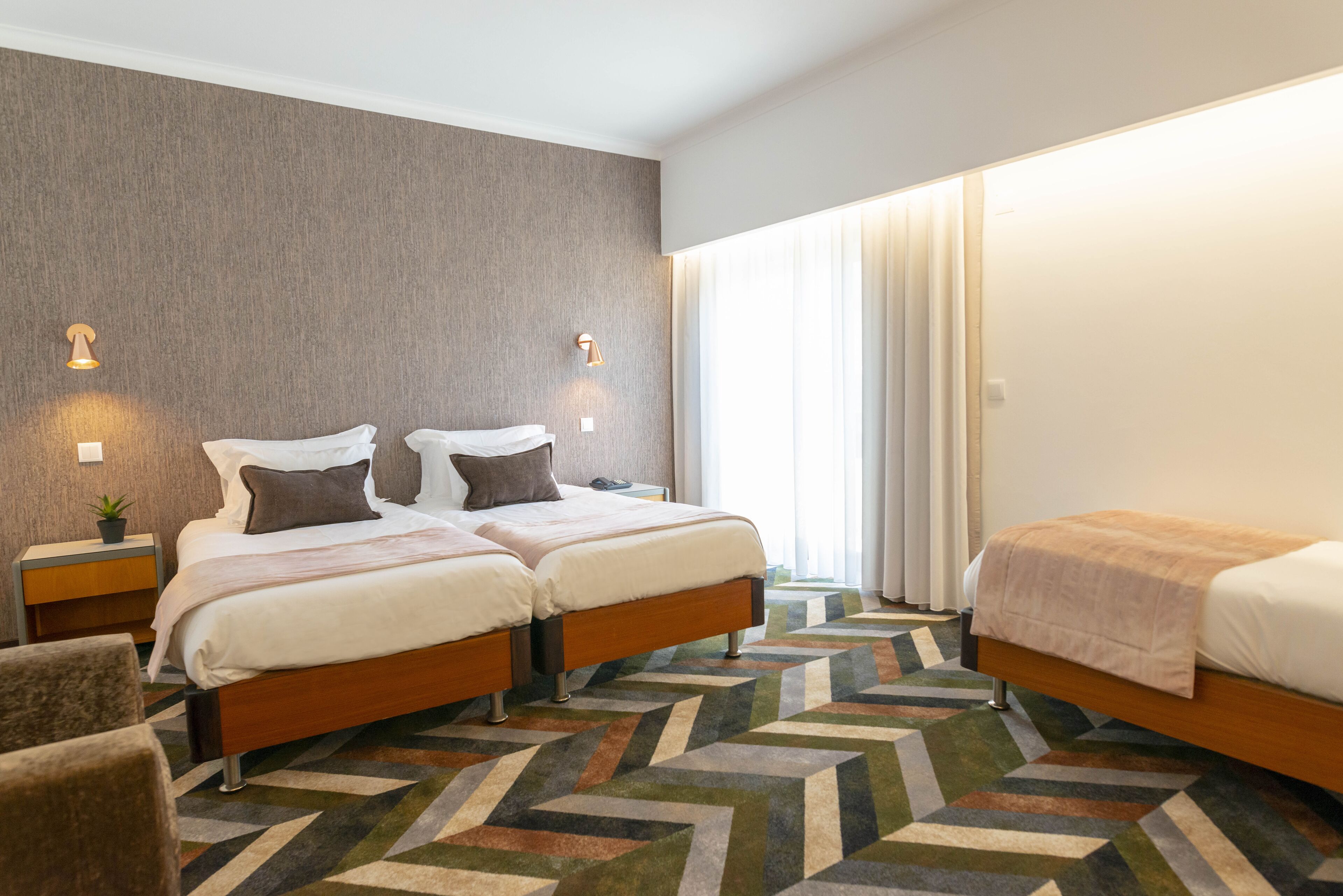 A hotel room with two beds, a patterned carpet, and a large window with curtains.