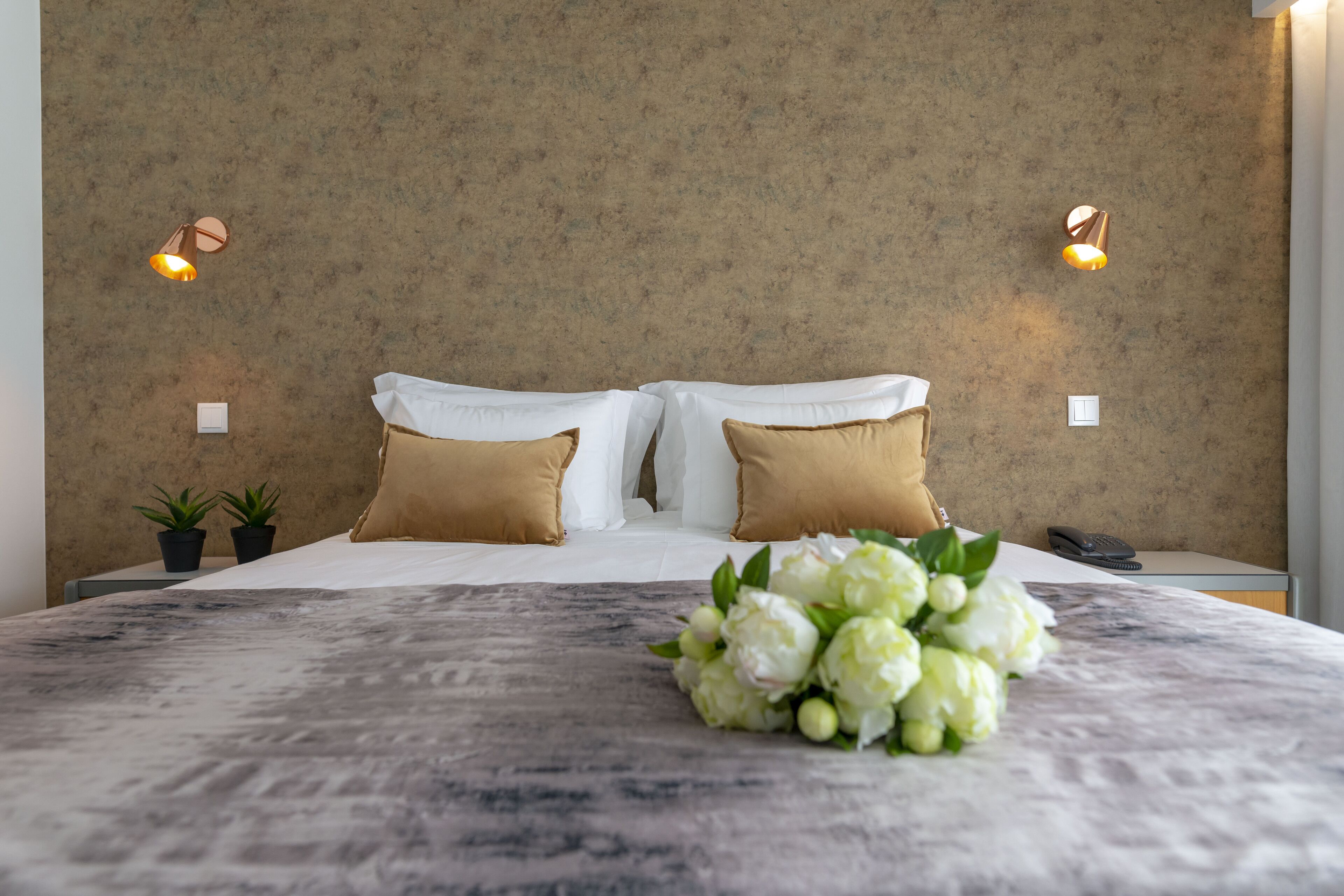 Cama bem arrumada com almofadas brancas e bege, um arranjo floral e duas pequenas plantas em vasos sobre a mesa de cabeceira.