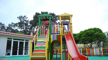 A colorful playground structure with slides and climbing elements, surrounded by a fence and trees.