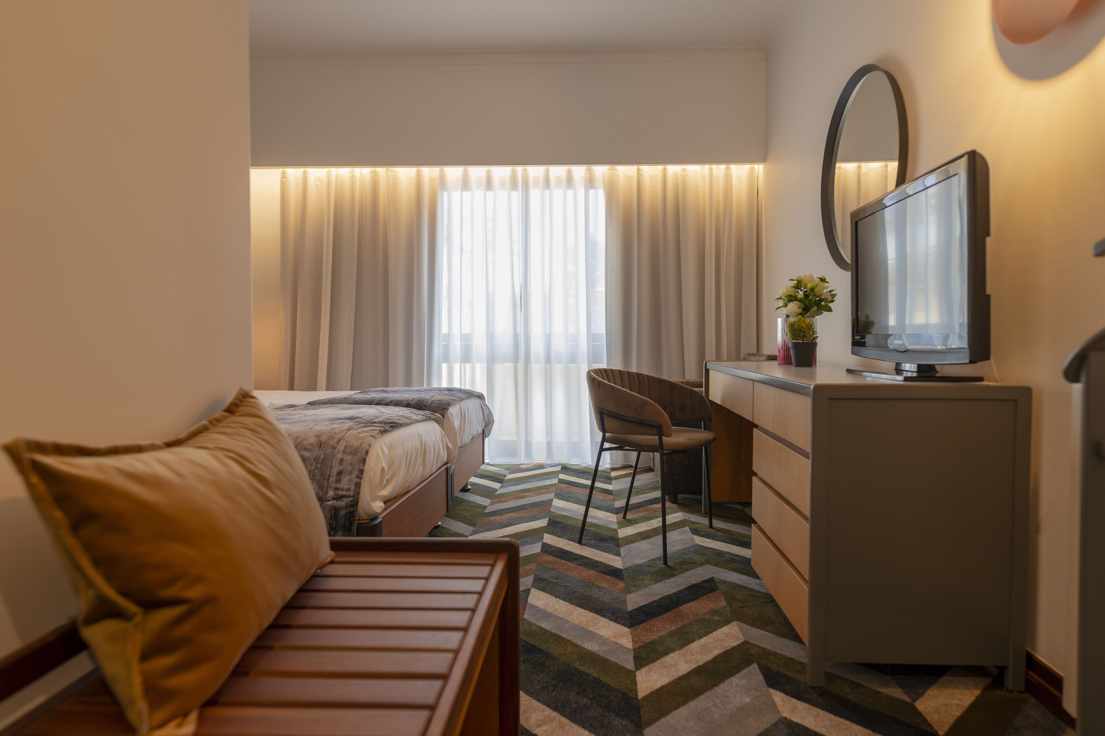 Una habitación de hotel con cama, escritorio, silla, televisor y ventana con cortinas.