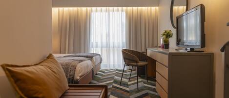 Una habitación de hotel con cama, escritorio, silla, televisor y ventana con cortinas.