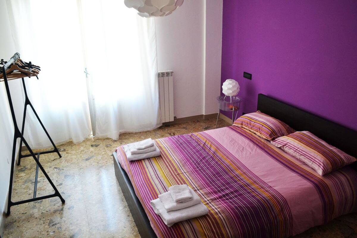 Een slaapkamer met een bed, een radiator, een kledingrek en een klein tafeltje met een decoratief object.