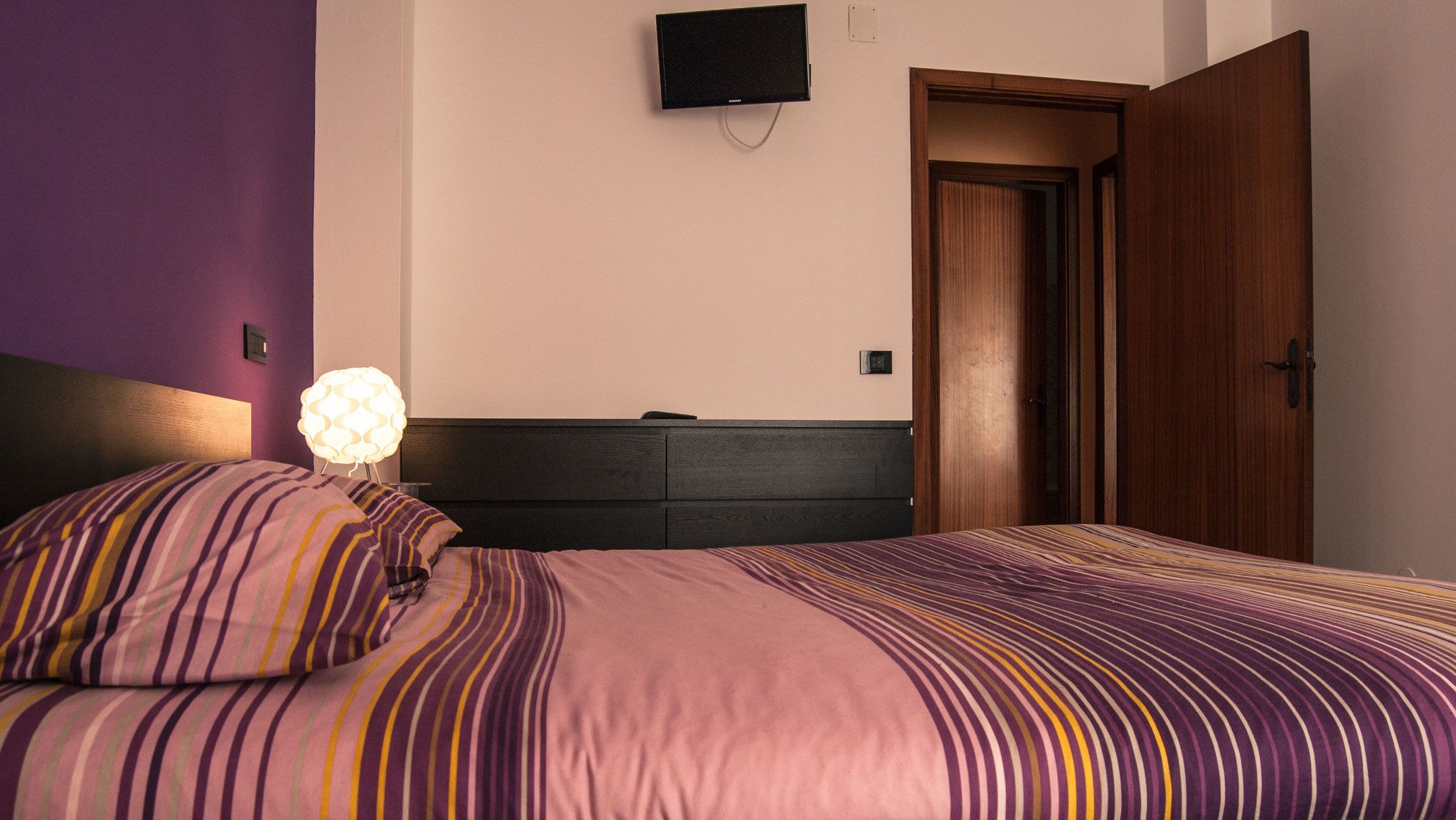 A bedroom with a bed, a bedside lamp, and a TV on the wall.