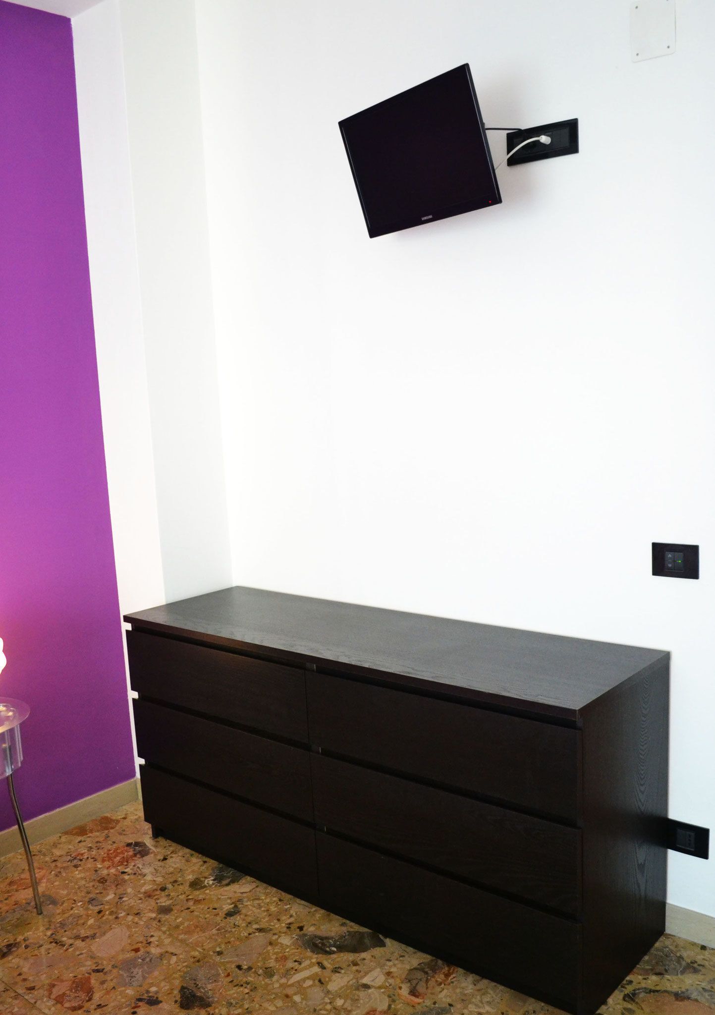 A room with a dark wooden dresser, a flat-screen TV mounted on the wall, and a purple accent wall.