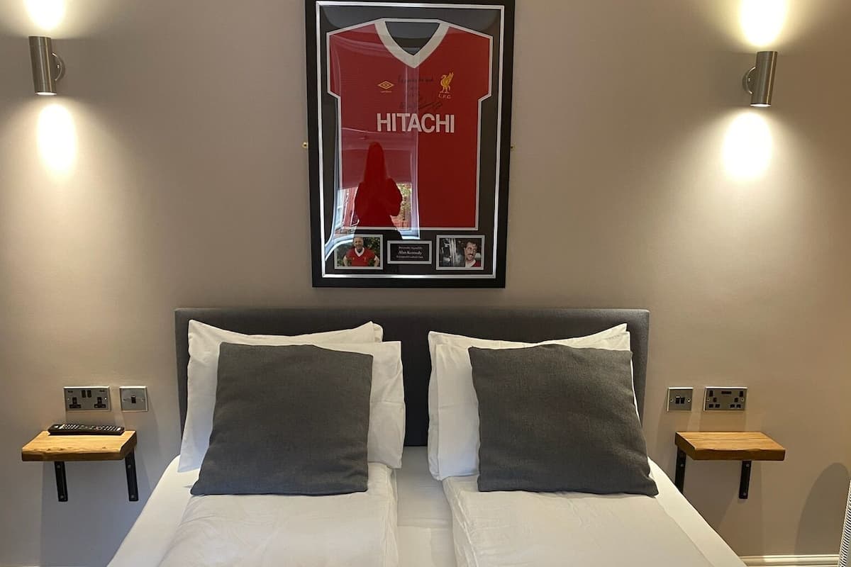 A double bed with white and grey bedding, two nightstands with lamps, a framed jersey on the wall, and folded towels on the bed.