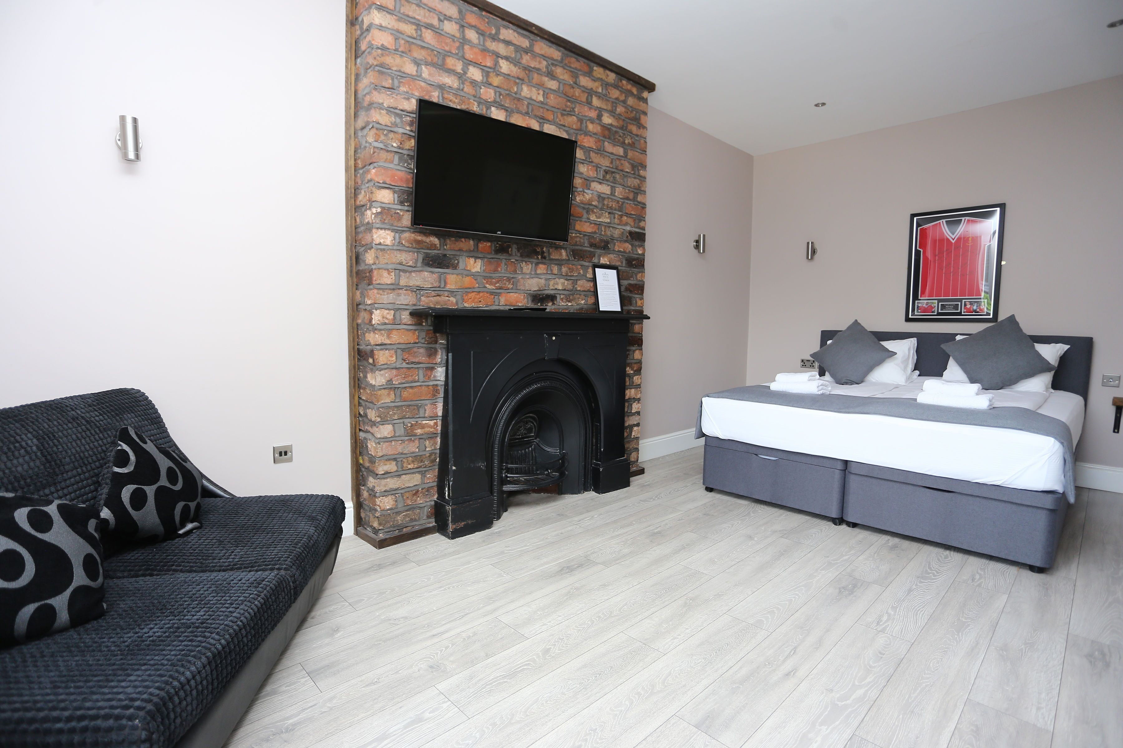 A modern bedroom with a brick fireplace, a flat-screen TV mounted above it, a bed with grey bedding, and a patterned sofa.
