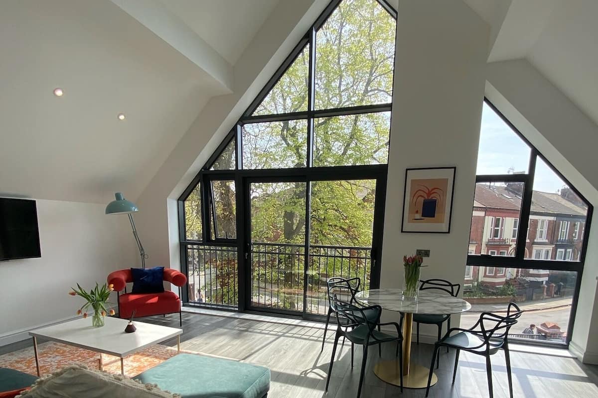A spacious loft with high ceilings, large windows, a dining area with a round table and chairs, a red sofa, and a TV mounted on the wall.