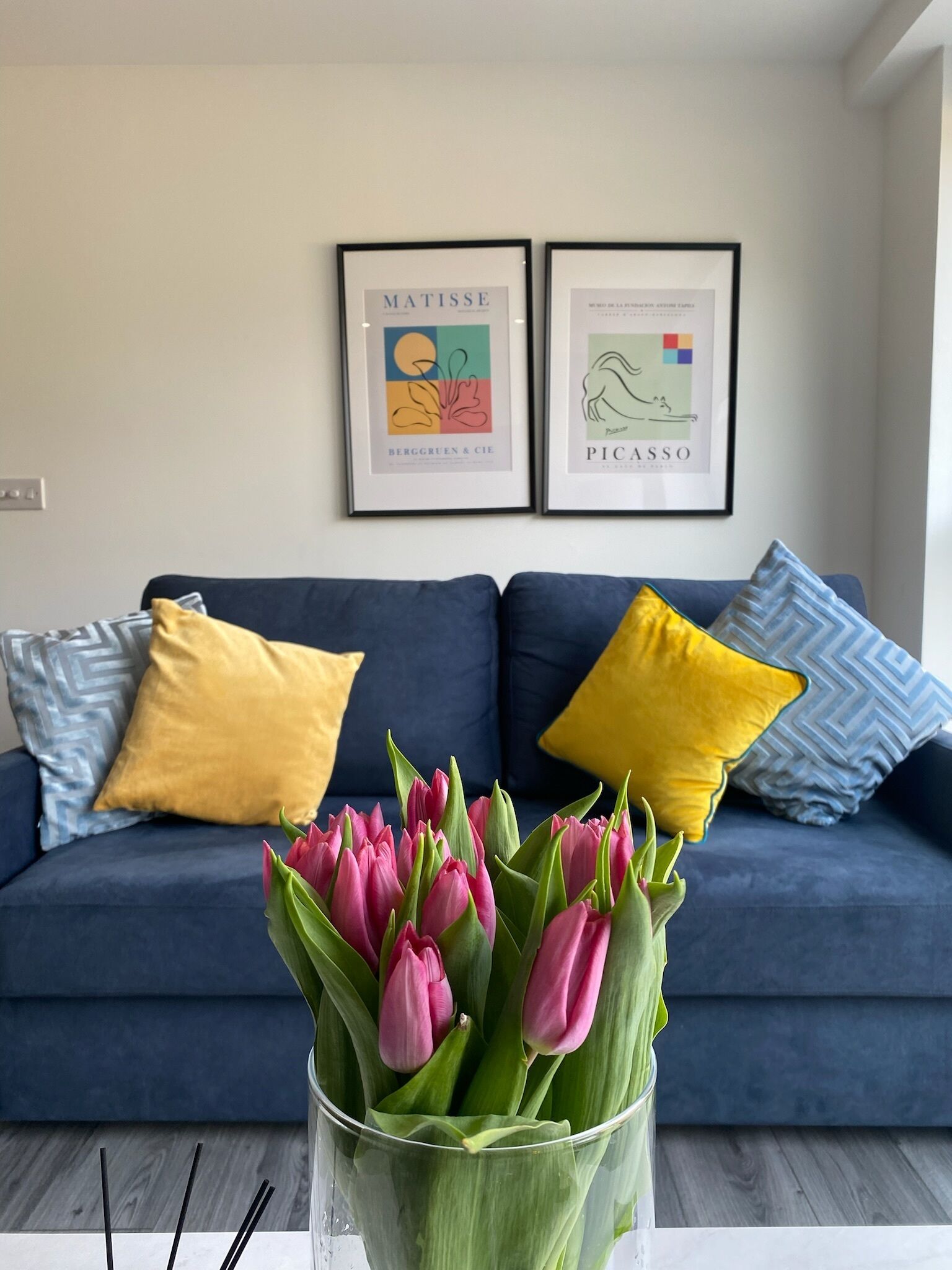 A living room with a blue sofa, yellow and patterned pillows, a vase of pink tulips, and two framed artworks on the wall.