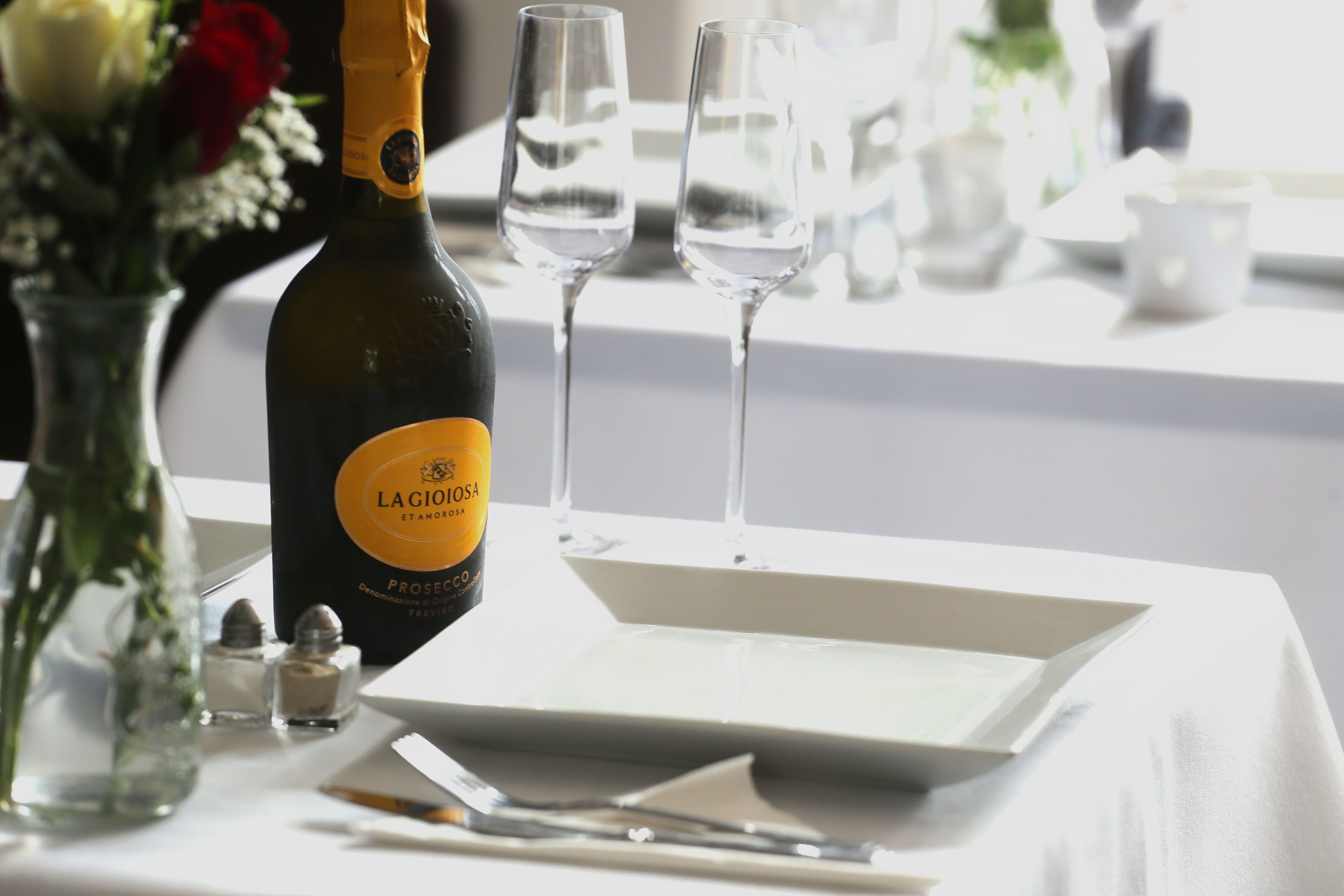 Una botella de La Cioiosa Prosecco, dos copas de vino, un plato blanco, un cuchillo, un tenedor, un pequeño jarrón con flores y saleros y pimenteros sobre una mesa.