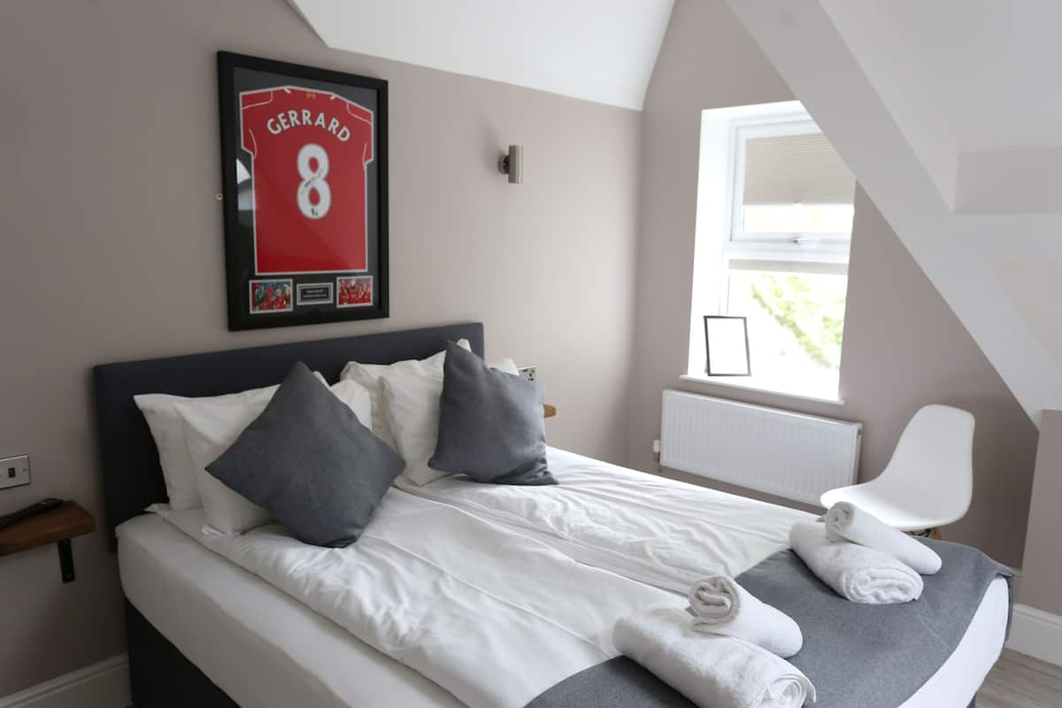 A neatly made bed with white linens and gray pillows, a framed sports jersey on the wall, a white chair, and a window with blinds.