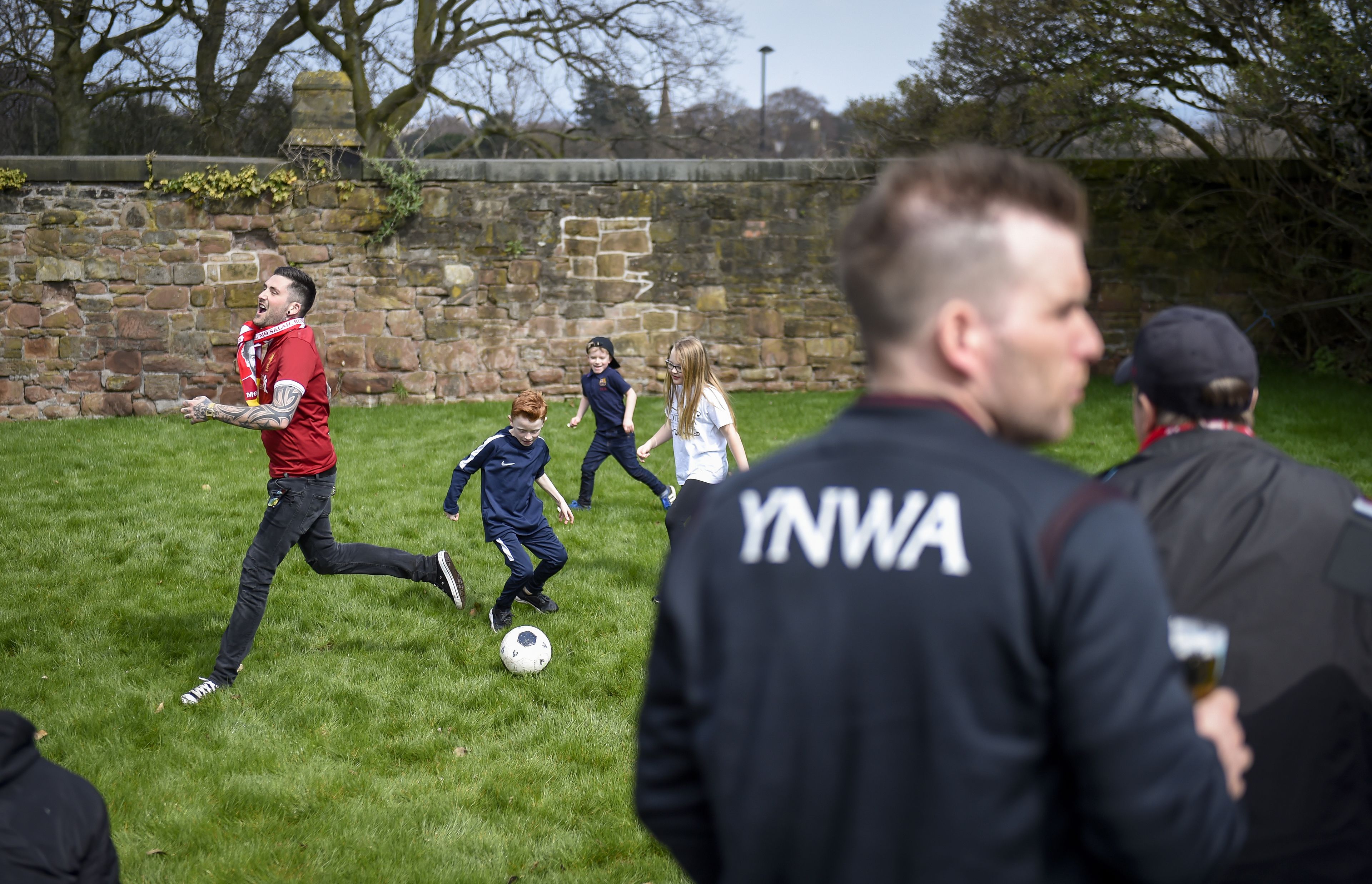 Un gruppo di persone gioca a calcio su un campo erboso, con un muro di pietra sullo sfondo.