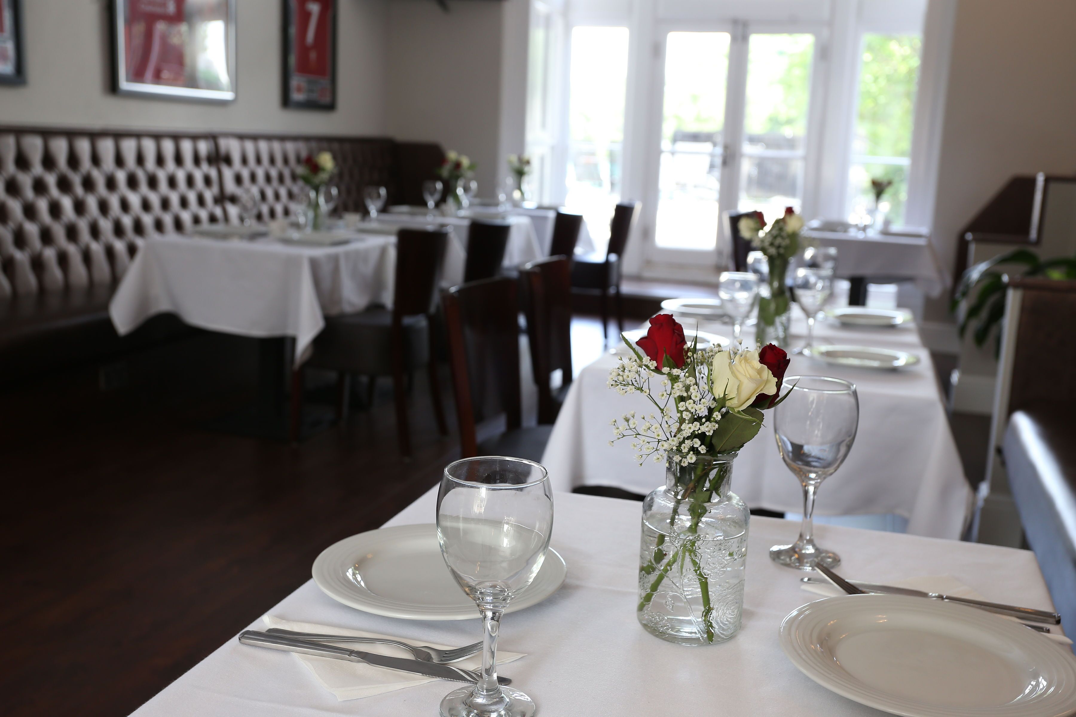 Un restaurante con manteles blancos, mesas dispuestas con platos, vasos y cubiertos. Hay jarrones con flores sobre las mesas.