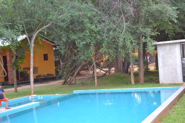 Una piscina con un niño sentado en el borde, rodeada de árboles y un edificio de fachada amarilla.