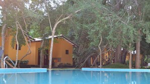 A swimming pool with a clear blue water surface, surrounded by trees and a building with a yellow facade. - Ayubowan Resorts (Avudangawa)