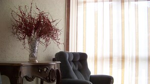 A room with a dark blue armchair, a wooden side table with a vase of red flowers, and a large window with vertical blinds. - Capri residence in Vicenza (vicenza)
