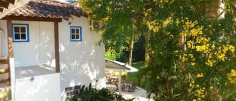 A white building with blue trim and a tiled roof, surrounded by lush greenery and yellow flowering trees.