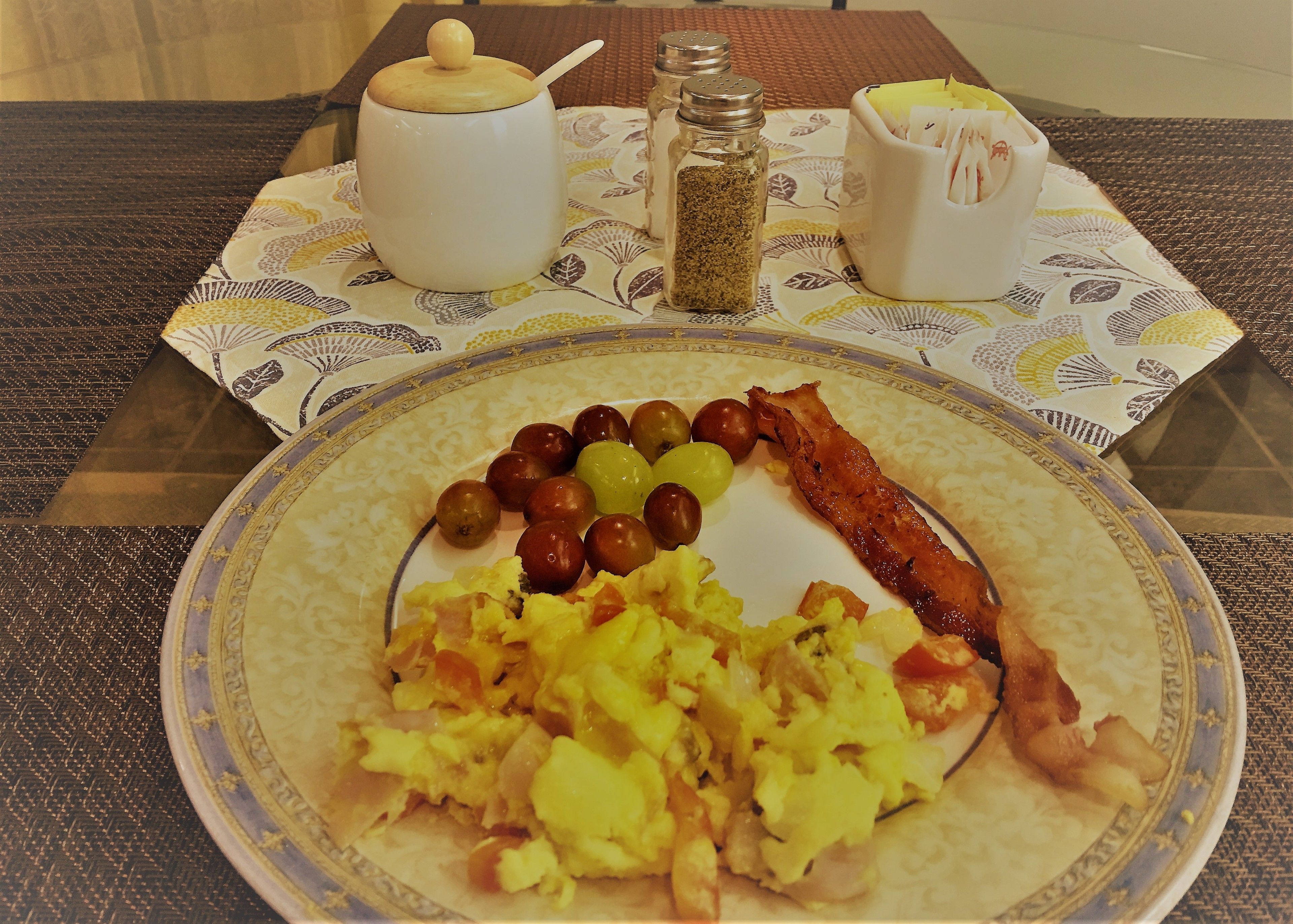 A breakfast plate with scrambled eggs, bacon, and olives on a patterned tablecloth.