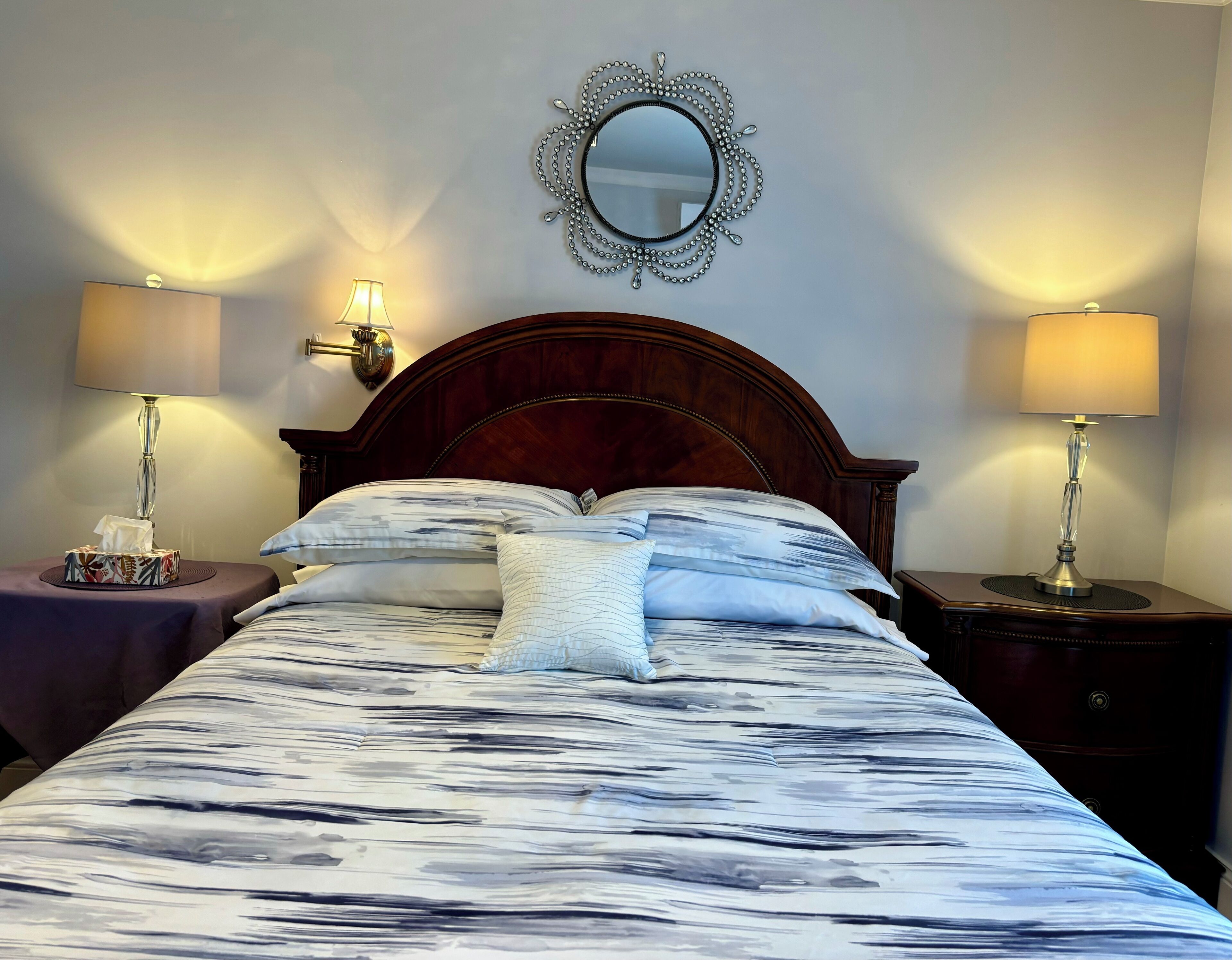 A neatly made bed with a marble-patterned bedspread, two bedside lamps, a decorative mirror, and a bedside table with a tissue box.