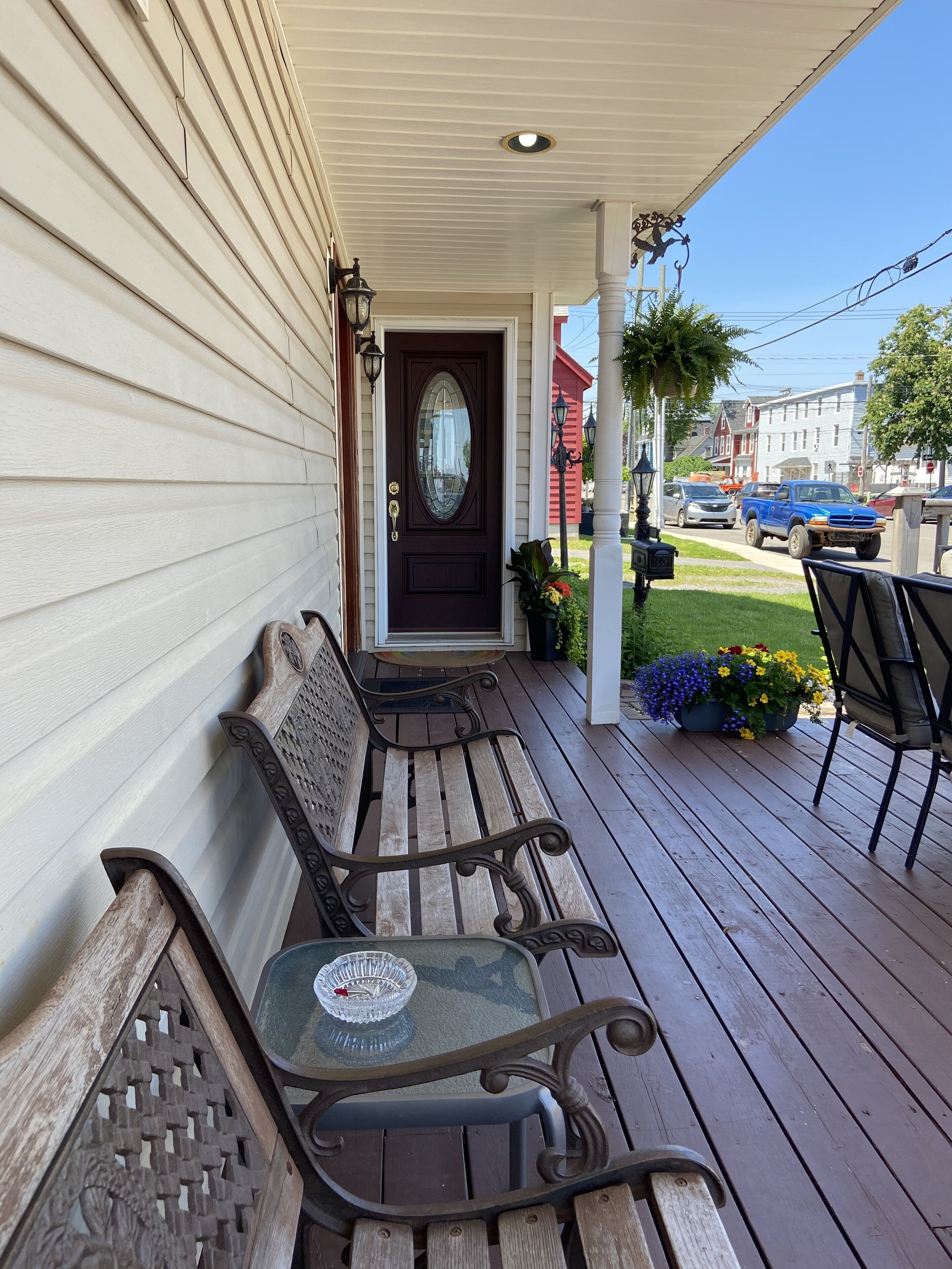 A wooden porch with two benches and a table, a dark front door, and a street view with cars and buildings in the background.