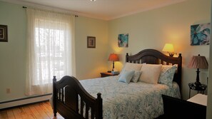 Ein Schlafzimmer mit einem hölzernen Bett, zwei Nachttischlampen, einem Fenster mit durchscheinenden Vorhängen und floralen Bildern an den Wänden.