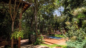 A wooden deck with a swimming pool, surrounded by lush greenery and a pergola structure.