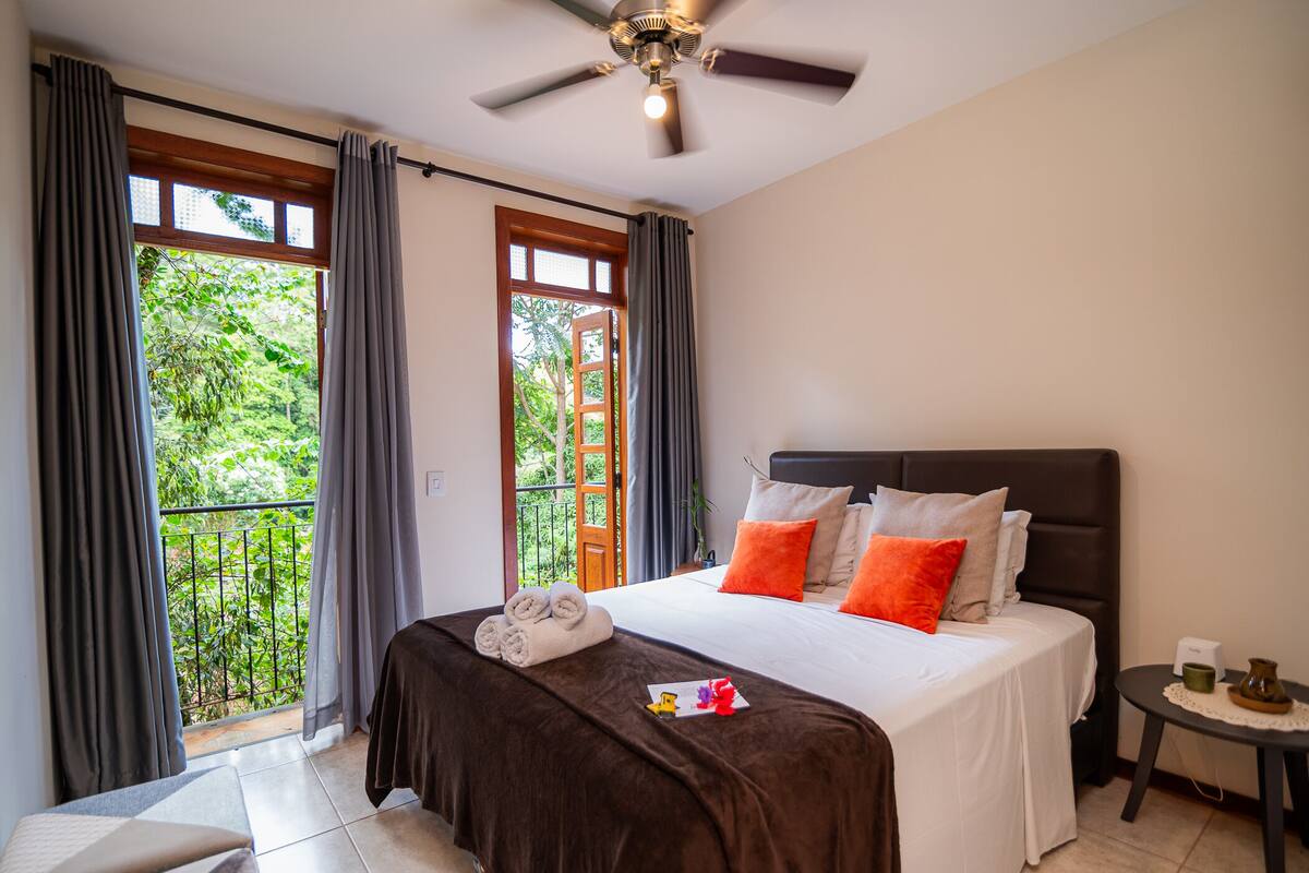 A hotel room with a bed, a small table, a ceiling fan, and a balcony with curtains.