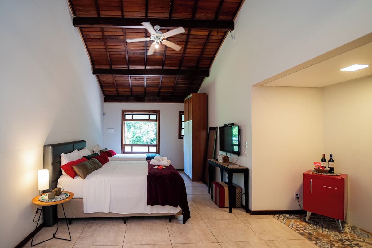 A hotel room with a bed, a TV, a ceiling fan, and a window with a view of trees.