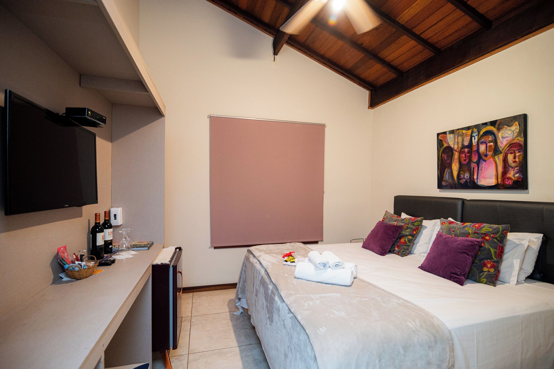 A hotel room with a bed, a flat-screen TV, a painting on the wall, and a window with a blind.